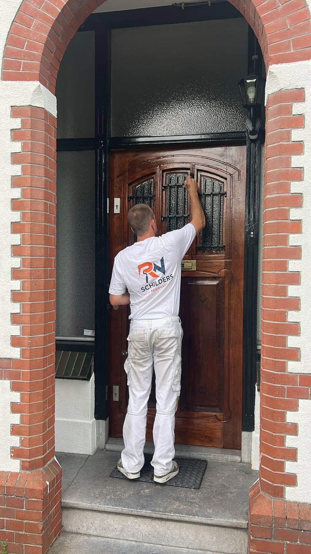 Schilder van RN Schilders werkt aan een houten voordeur in een bakstenen entree